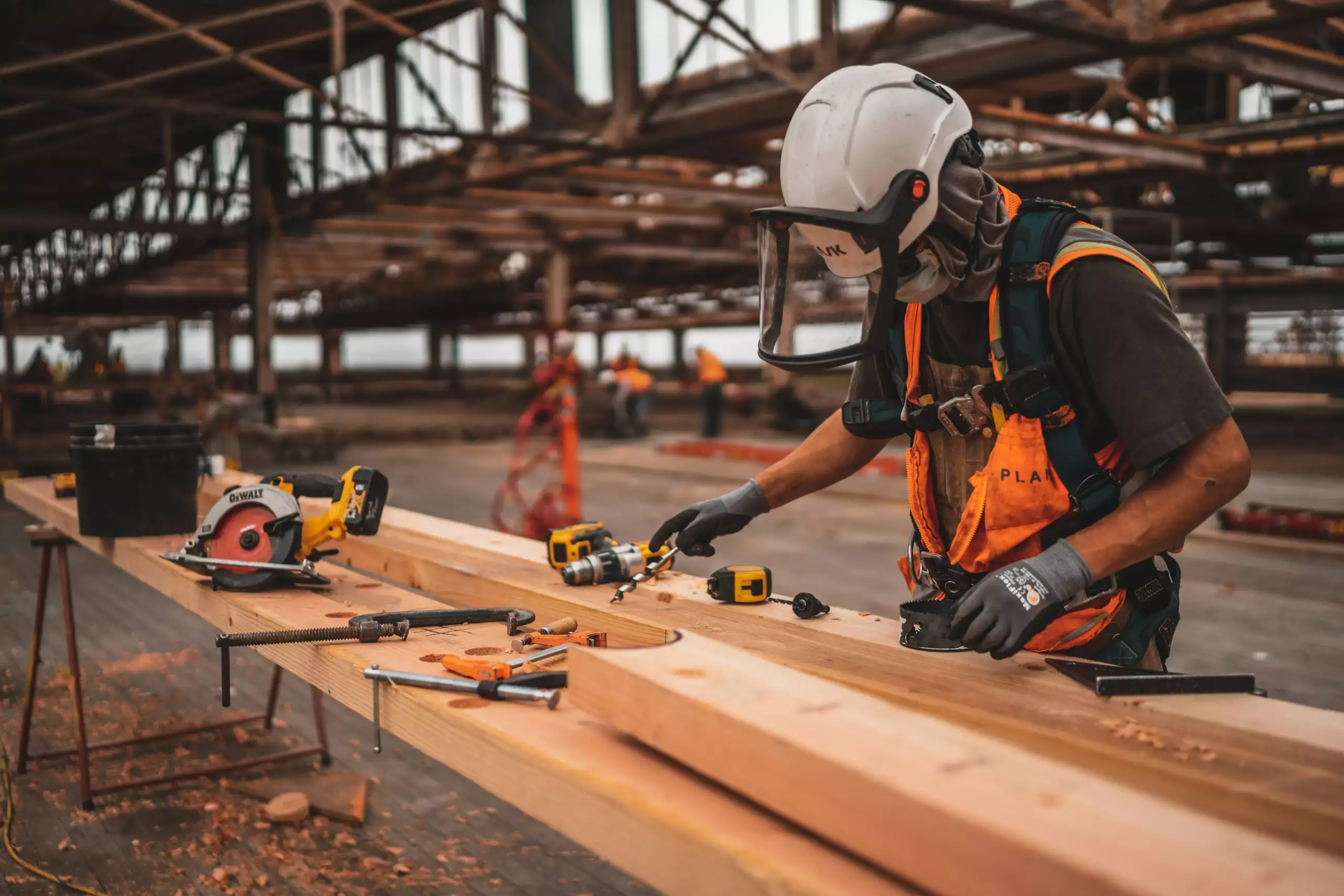 A man in orange and black vest wearing white helmet holding yellow and black power tool